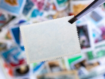 Blank postage stamp in tweezers against blurred background collection of multicolored postage stamps